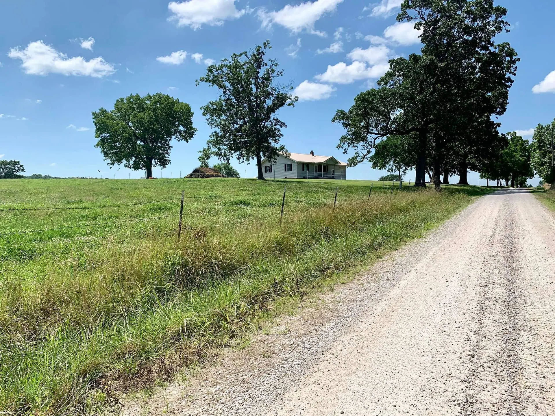 Gravel farm road running past open pasture toward the farmhouse under a wide blue Missouri sky