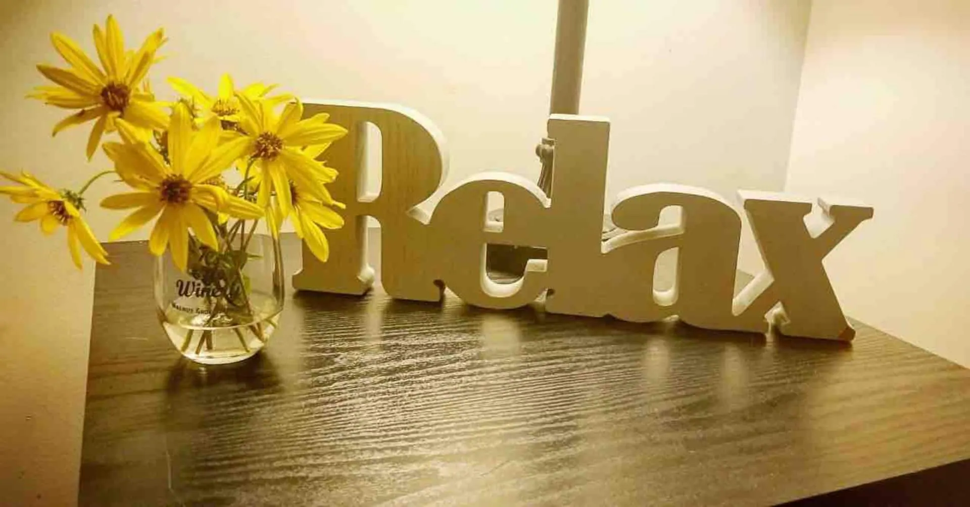 A wooden "Relax" sign beside a jar of cut sunflowers on the table